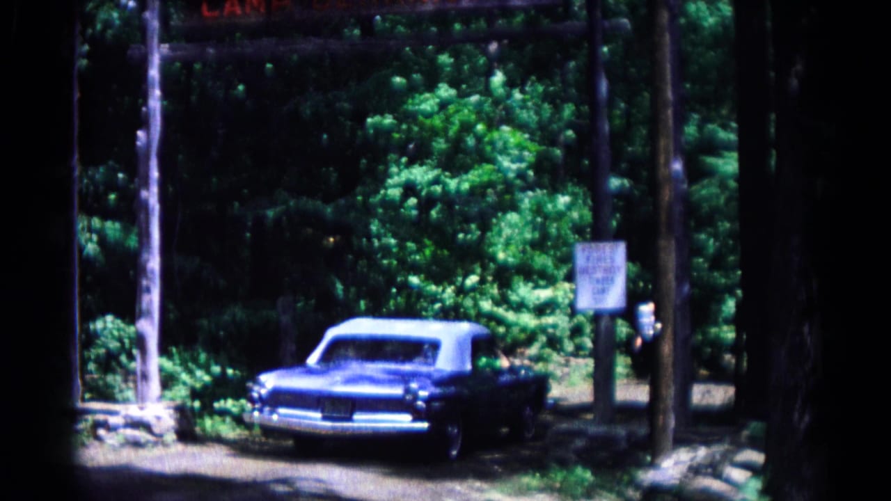 The vintage blue convertible moves along the dirt entrance road beneath the Camp Ockanickon sign in the wooded camp setting.