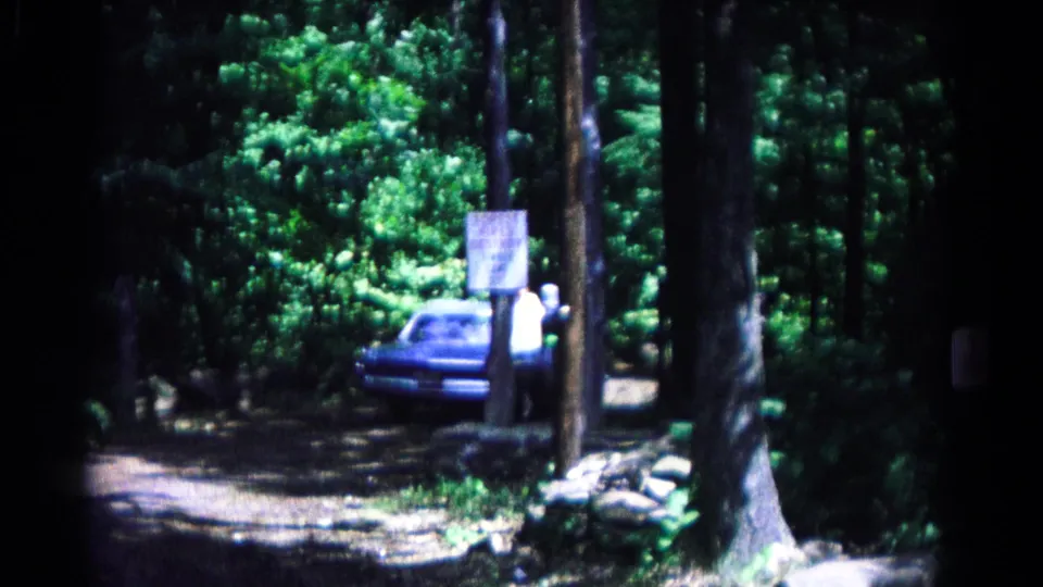 From behind, the blue convertible proceeds down the shaded dirt road past the camp entrance posts and forest edge.