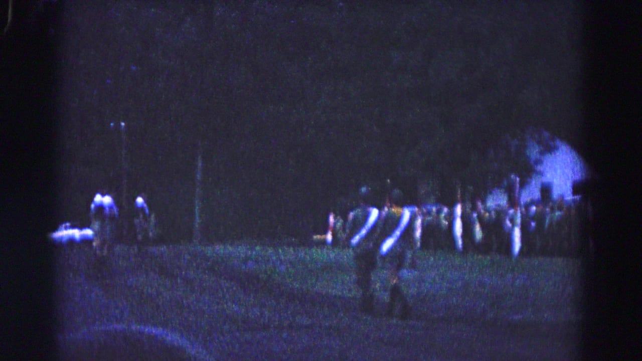 Uniformed men march across a dark grassy field with spectators and trees in the background.