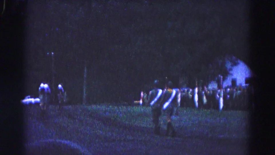 1966 Hagerstown home-movie frame showing uniformed men crossing a dim grassy field near spectators and trees.