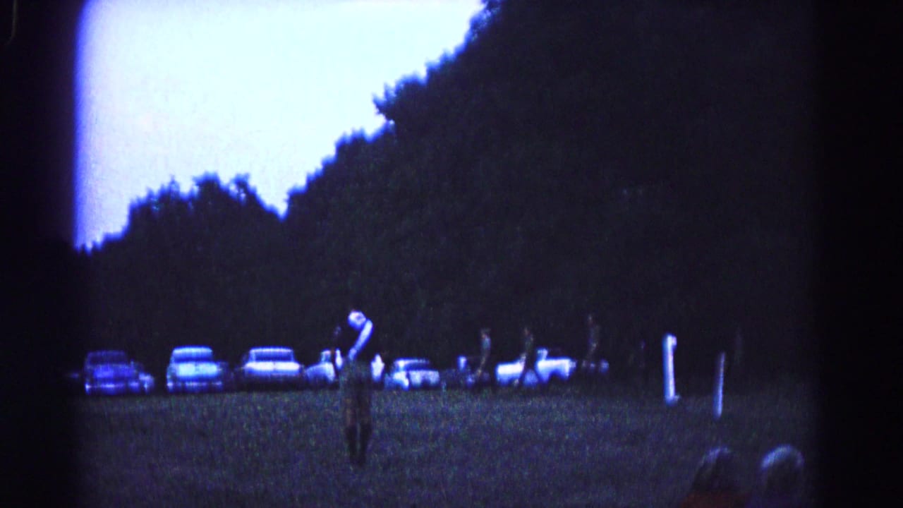 A uniformed man stands on grass with parked cars and dark trees behind him.