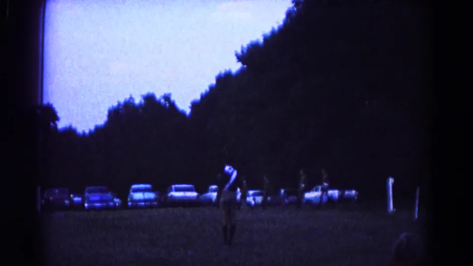 Home-movie frame from Hagerstown in 1966 showing a uniformed figure on grass with mid-century cars and trees in the background.