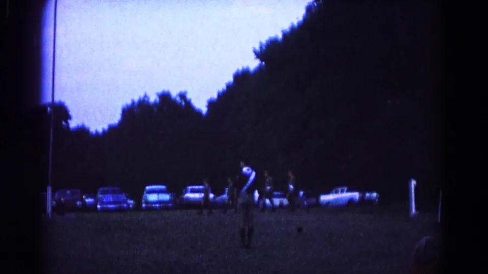 1966 Hagerstown frame showing uniformed participants on a grassy field, with parked cars and spectators along the tree line.