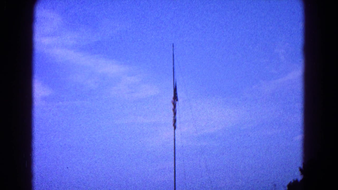 Distant American flag on a pole in 1966 Hagerstown home-movie footage, framed by blue sky and faint clouds.
