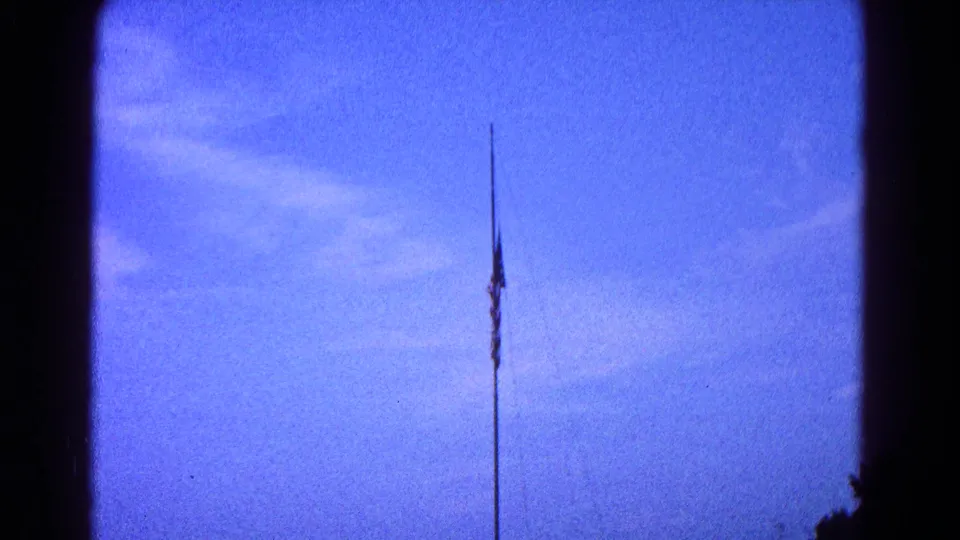 Distant American flag on a pole in 1966 Hagerstown home-movie footage, framed by blue sky and faint clouds.