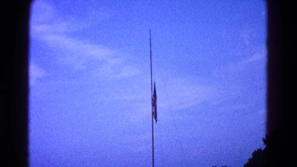 Final 1966 home-movie frame of the American flag lowered on a pole, with blue sky and dark edge silhouettes visible.