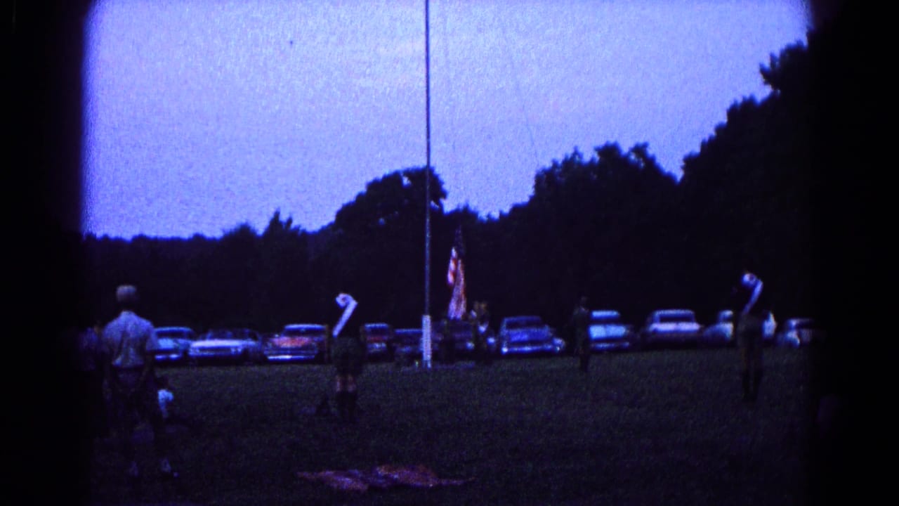 Scout-like figures stand near a flagpole on a dusk field with parked cars and trees behind them.