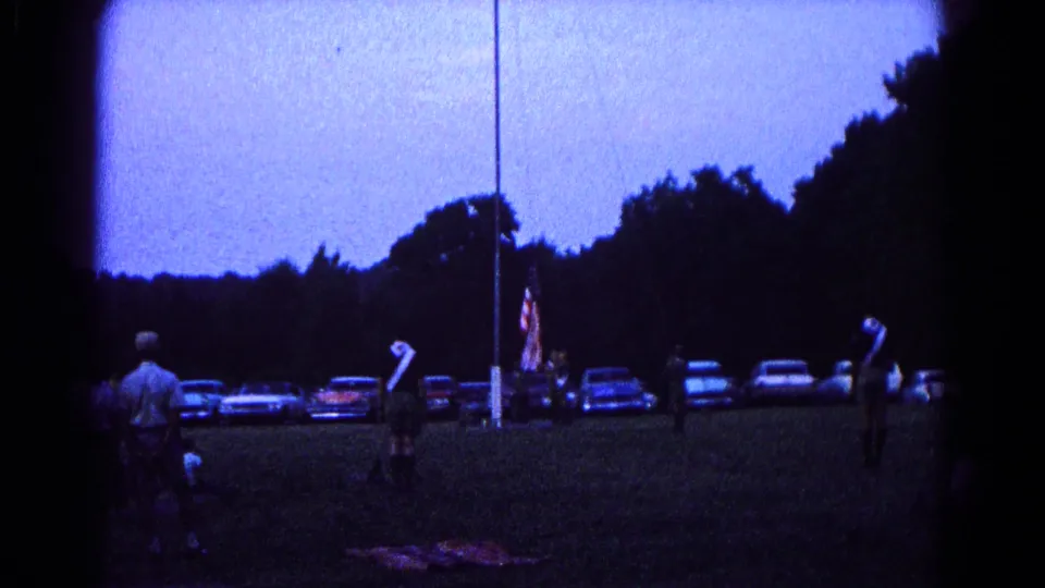 1966 Hagerstown home-movie frame showing scout-like participants near a flagpole as cars line the field at dusk.