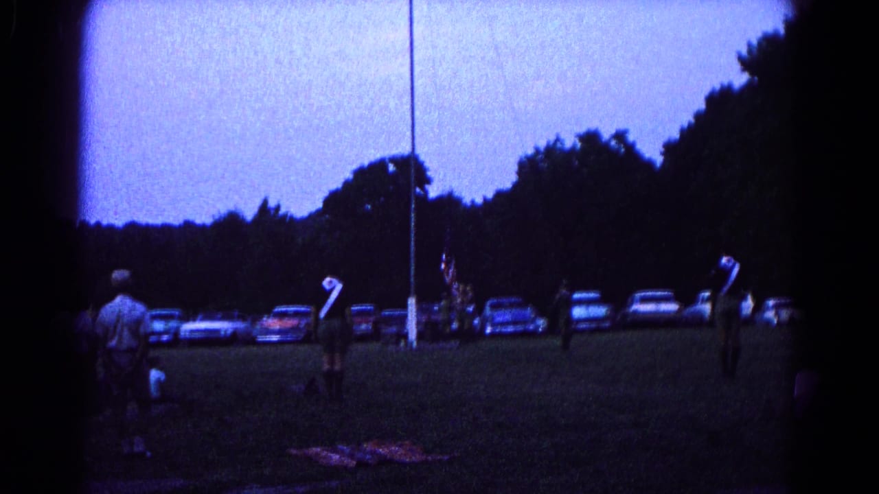 A group stands around a flagpole on a darkening field with a row of cars in the background.