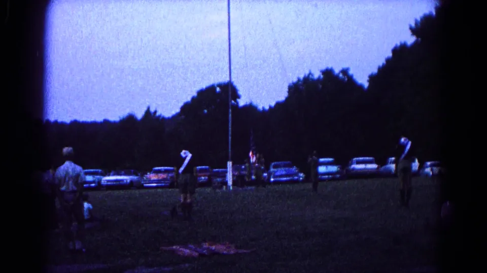 Twilight 8mm frame from Hagerstown showing a flagpole ceremony with distant participants and mid-1960s cars nearby.