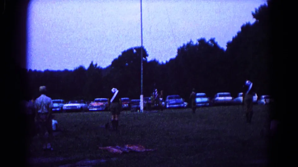 End frame of a 1966 Hagerstown flag-lowering scene, with scout-like figures, parked cars, and a dark tree line at dusk.