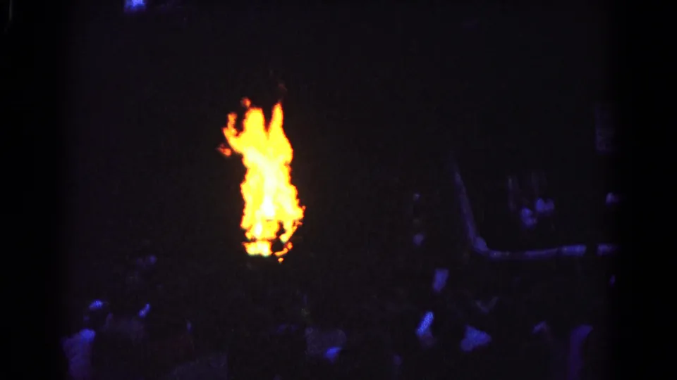 1966 Hagerstown home-movie frame showing a bright outdoor fire at night with tree silhouettes and people gathered in darkness.
