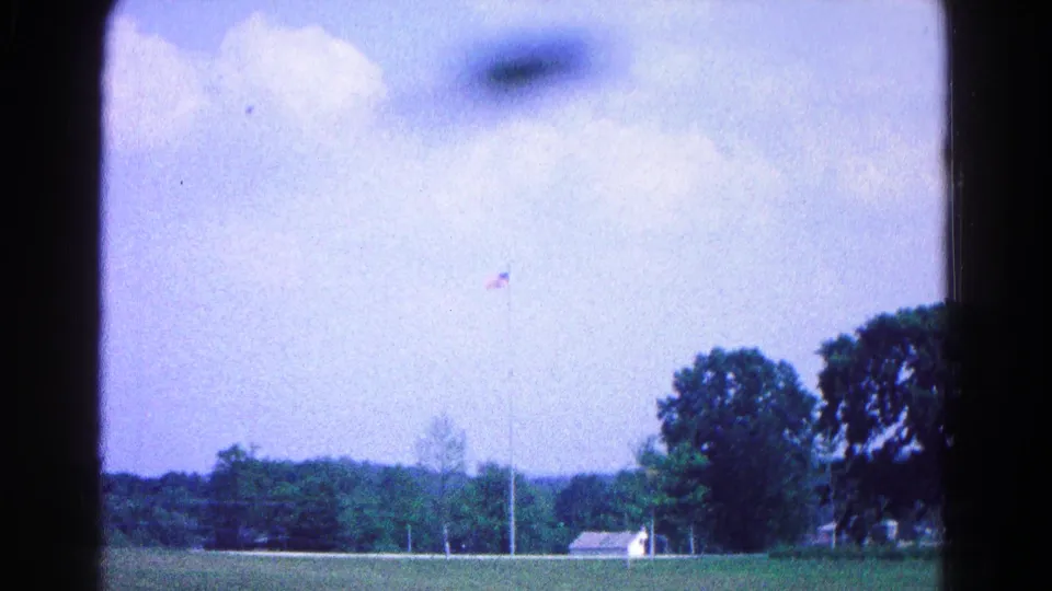 1966 8mm frame of a Hagerstown memorial or museum lawn, with a flagpole, trees, and small building visible in soft archival color.