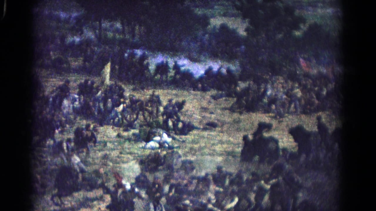 Groups of reenactors gather on a field with flags and several figures lying on the ground.