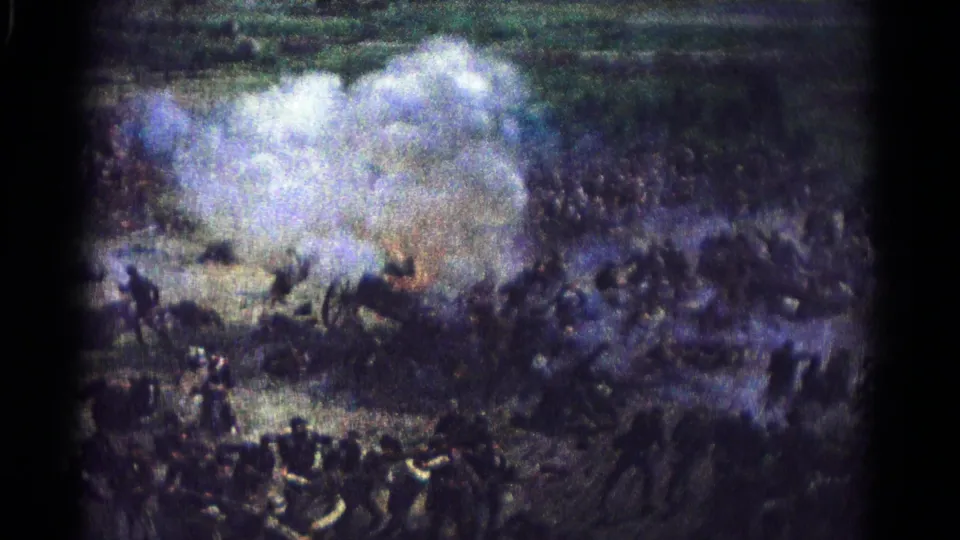 The reenactment field is partly obscured by cannon smoke, with uniformed participants and prone figures visible through the haze.
