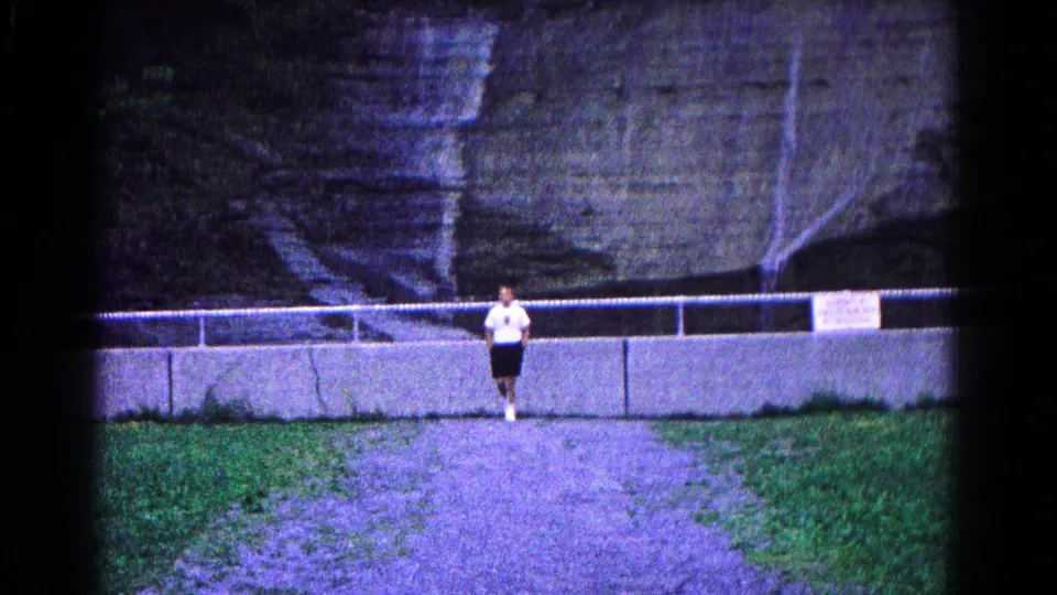 In Hagerstown, Maryland, 1966, a young man in a white shirt and dark shorts stands near a concrete barrier below layered rock.