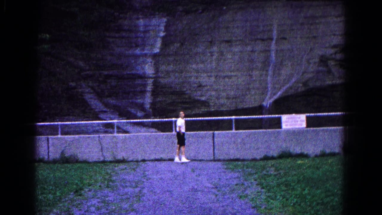 The man walks along the park path in Hagerstown, with a railing, posted sign, and layered stone cliff behind him.