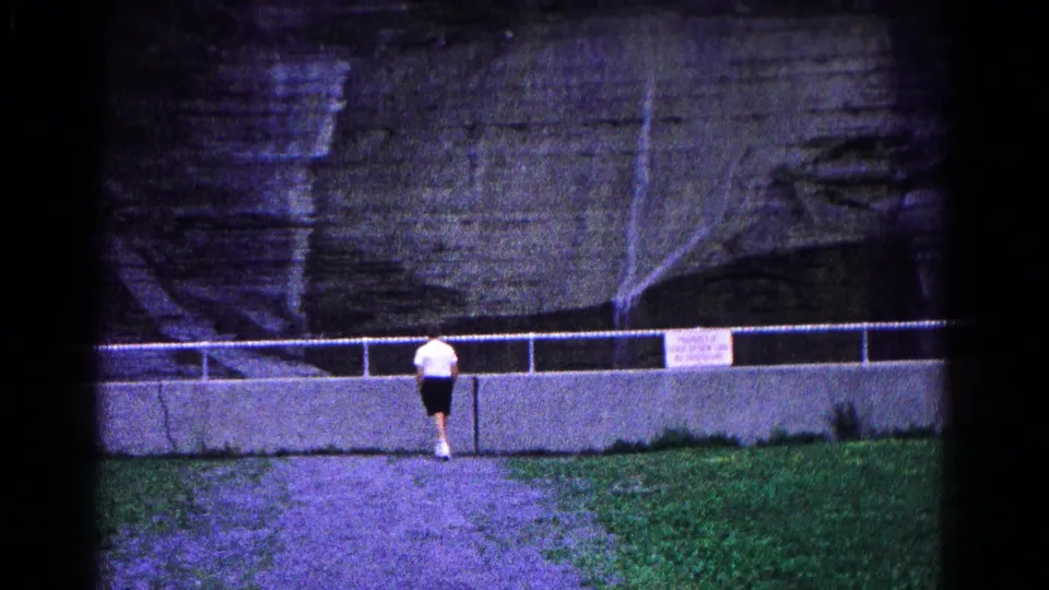 The young man turns away from the camera on the gravel path near the low wall and railing below the rock formation.