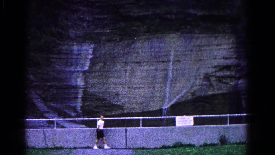 A young man in shorts walks left along the park barrier, framed against grass and layered rock in the 1966 Hagerstown footage.