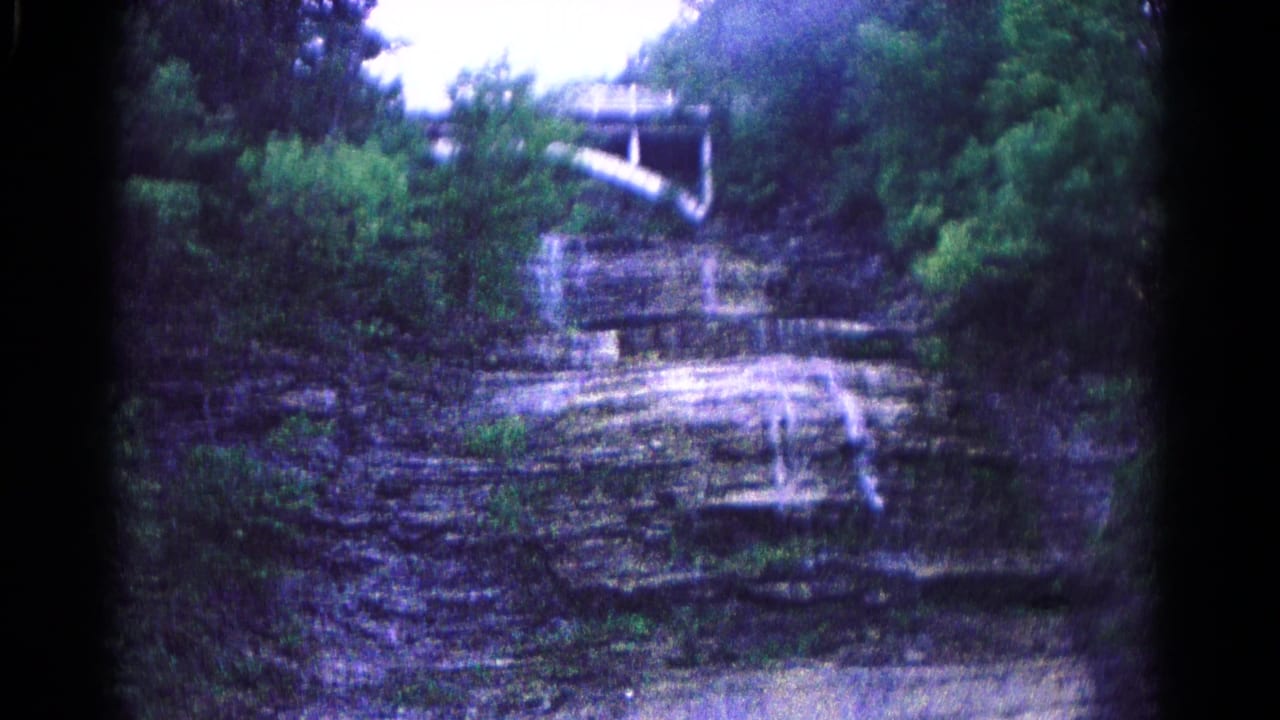 Wooded cliff face with water trickling down layered rock and a white structure above.