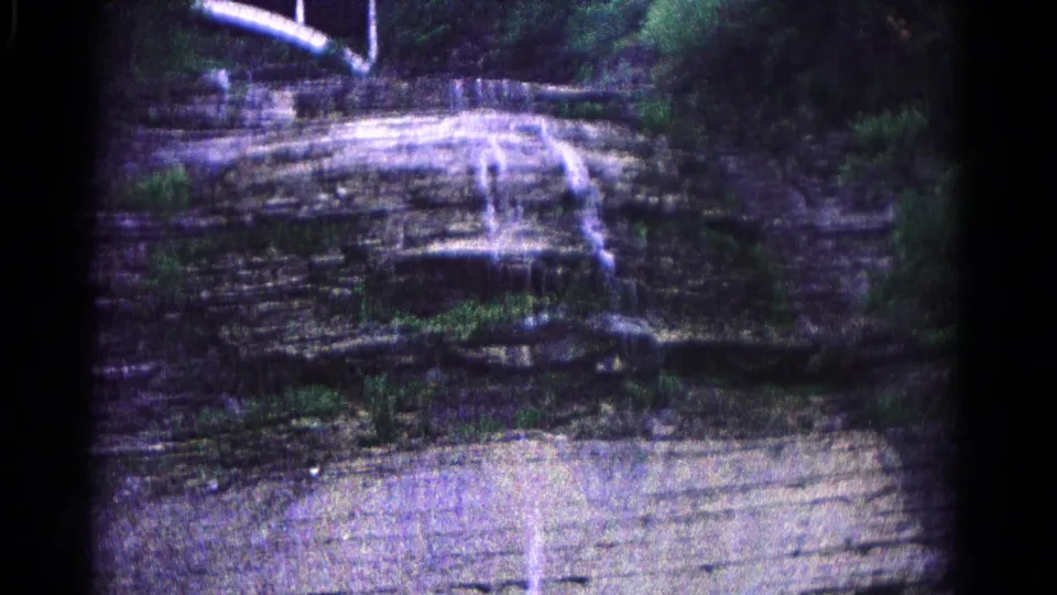 1966 8mm frame of a wooded Hagerstown-area cliff, with water dropping over layered rock and dense greenery along the sides.