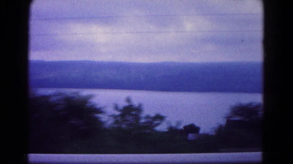 1966 8mm moving shot near Hagerstown with blurred roadside trees in the foreground, a calm water surface, and hazy wooded hills beyond.
