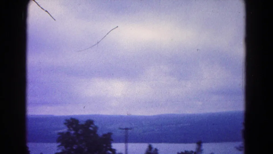 Soft 1966 8mm frame showing water, hills, a low tree line, and a small utility pole beneath a cloudy sky.