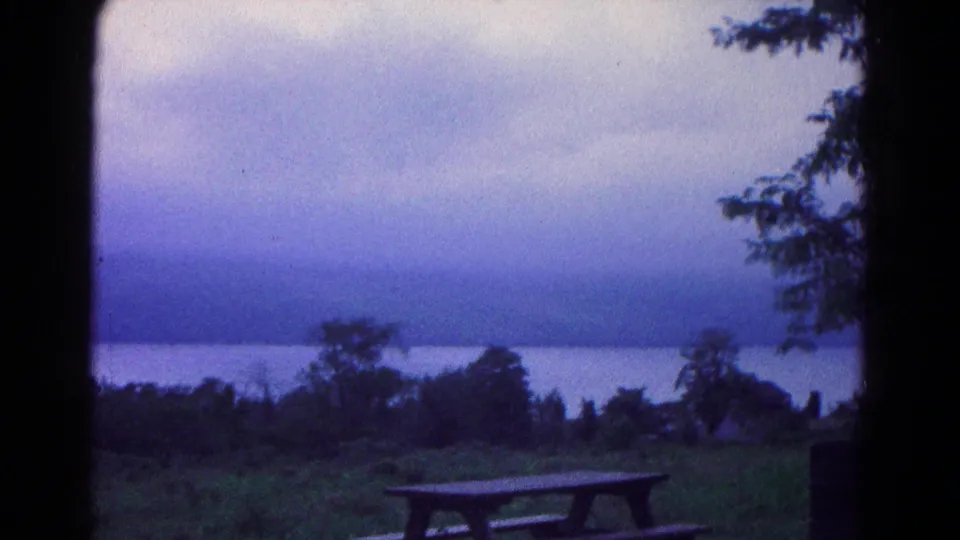 1966 Hagerstown-area home-movie frame with a picnic table at a grassy overlook above water and distant hills.