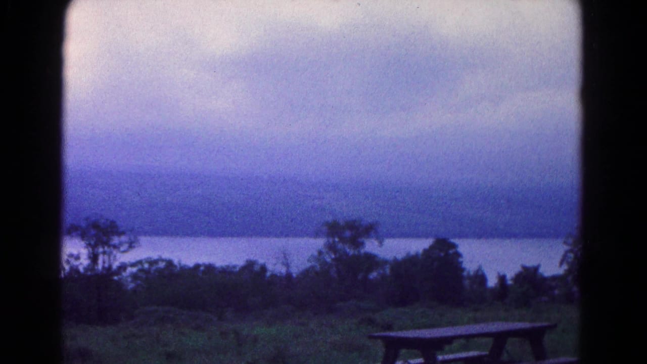 Overcast sky above water, distant hills, trees, grass, and a picnic table.