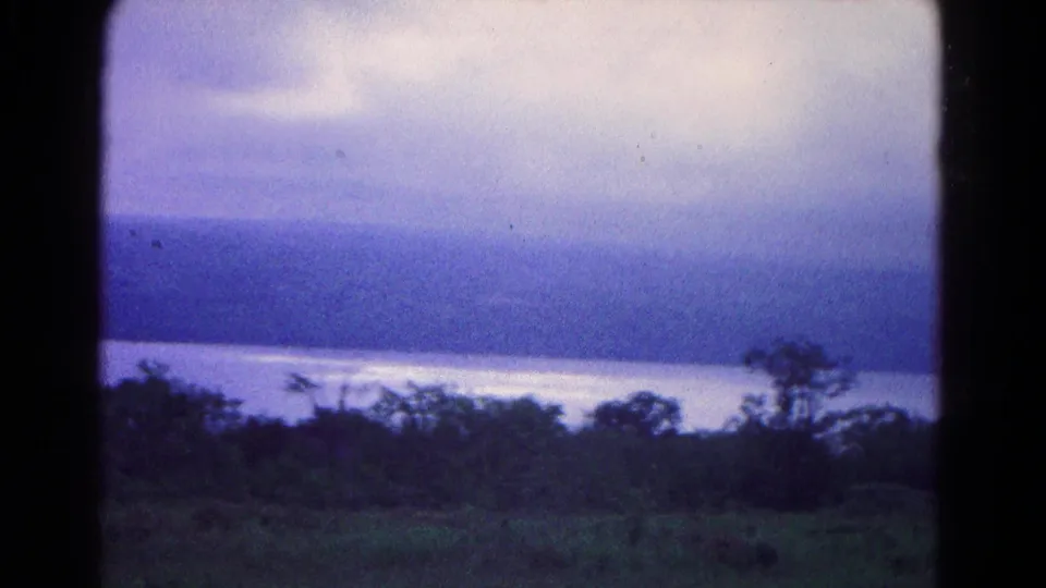 1966 8mm Hagerstown-area lakeside view with muted clouds, reflective water, wooded hills, grassy foreground, and a partial picnic table.