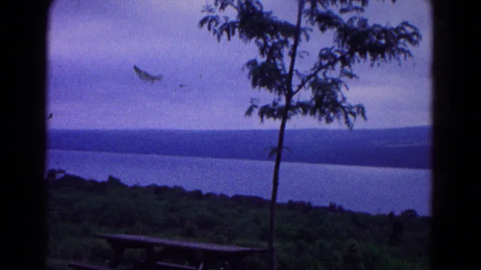 1966 home-movie view near Hagerstown with a picnic table, slender tree, water, wooded ridge, and muted overcast sky.