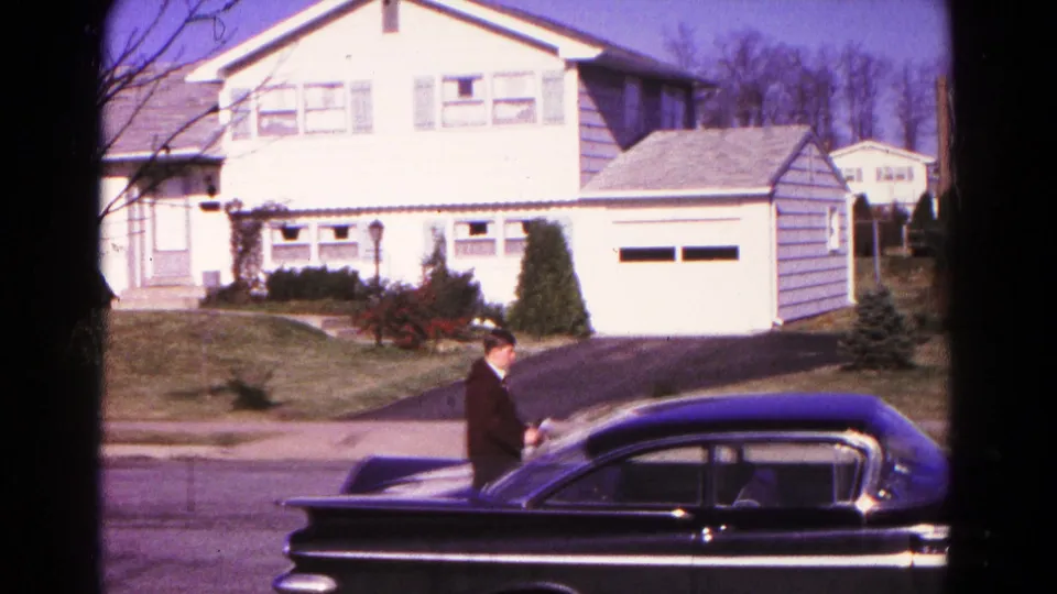 1966 Hagerstown suburban street frame with a white house, garage, dark sedan, driveway, and suited adult near the car.