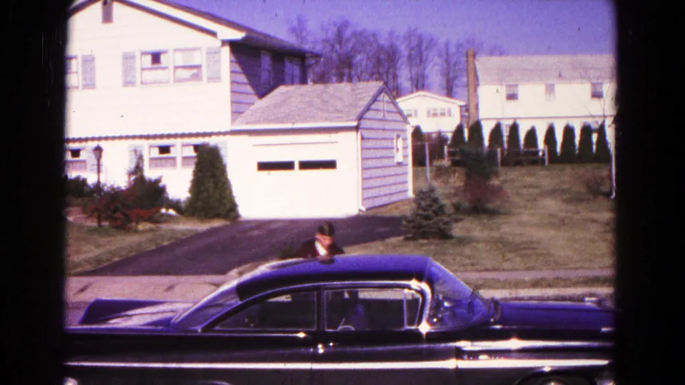 1966 Hagerstown home-movie frame showing a dark sedan, white suburban houses, lawn, and an adult partly visible by the car.