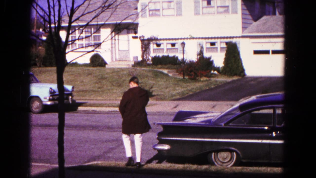 Man in a dark coat walks near a black sedan on a suburban street.