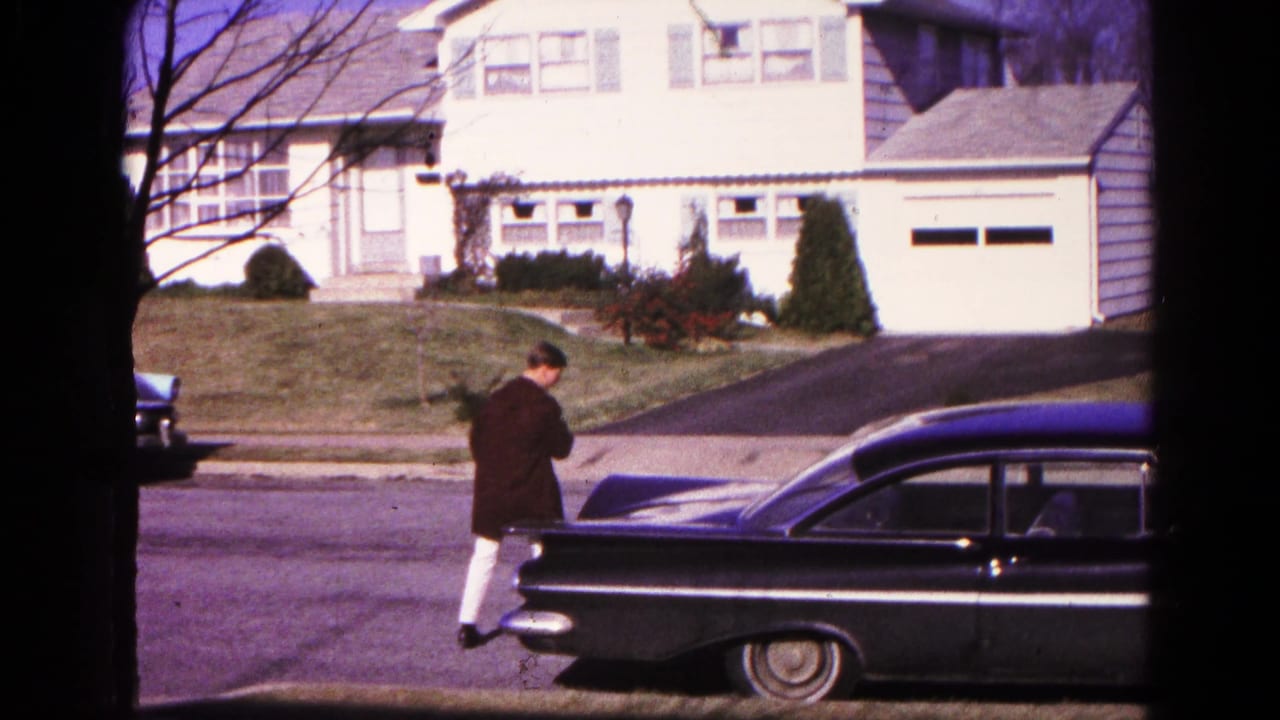 The man approaches the rear of a black sedan on a residential street in the supplied 1966 Hagerstown home-movie footage.