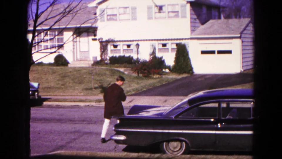 The man approaches the rear of a black sedan on a residential street in the supplied 1966 Hagerstown home-movie footage.