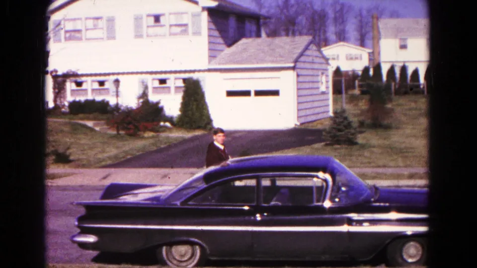 A black mid-century sedan passes across the foreground while the man appears near its driver side in the 1966 Hagerstown street scene.