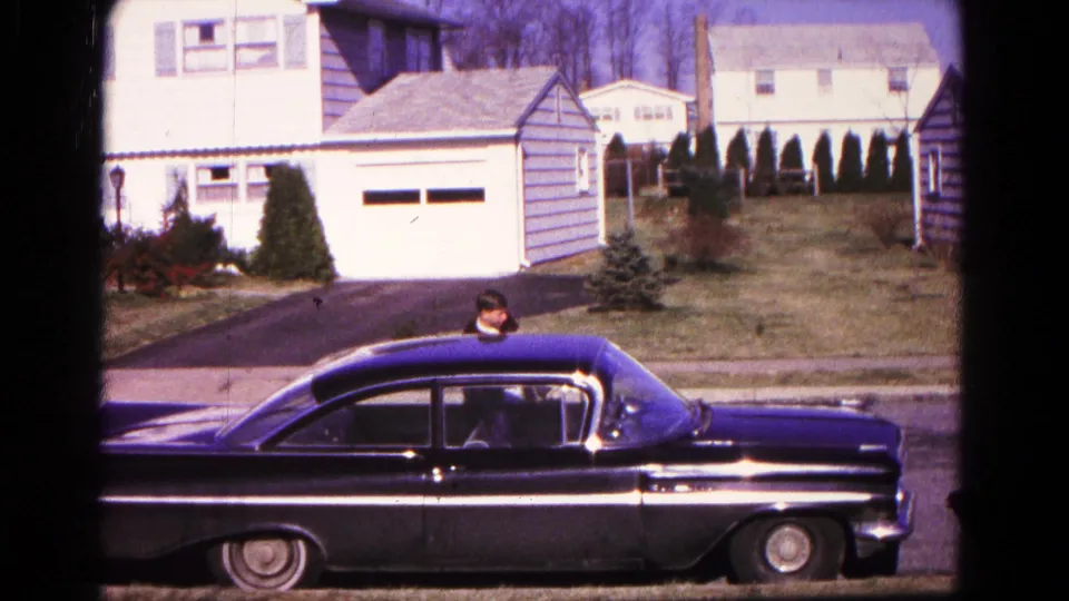 The black sedan sits along the curb in 1966 Hagerstown, with the man partly visible behind or beside it and suburban houses beyond.