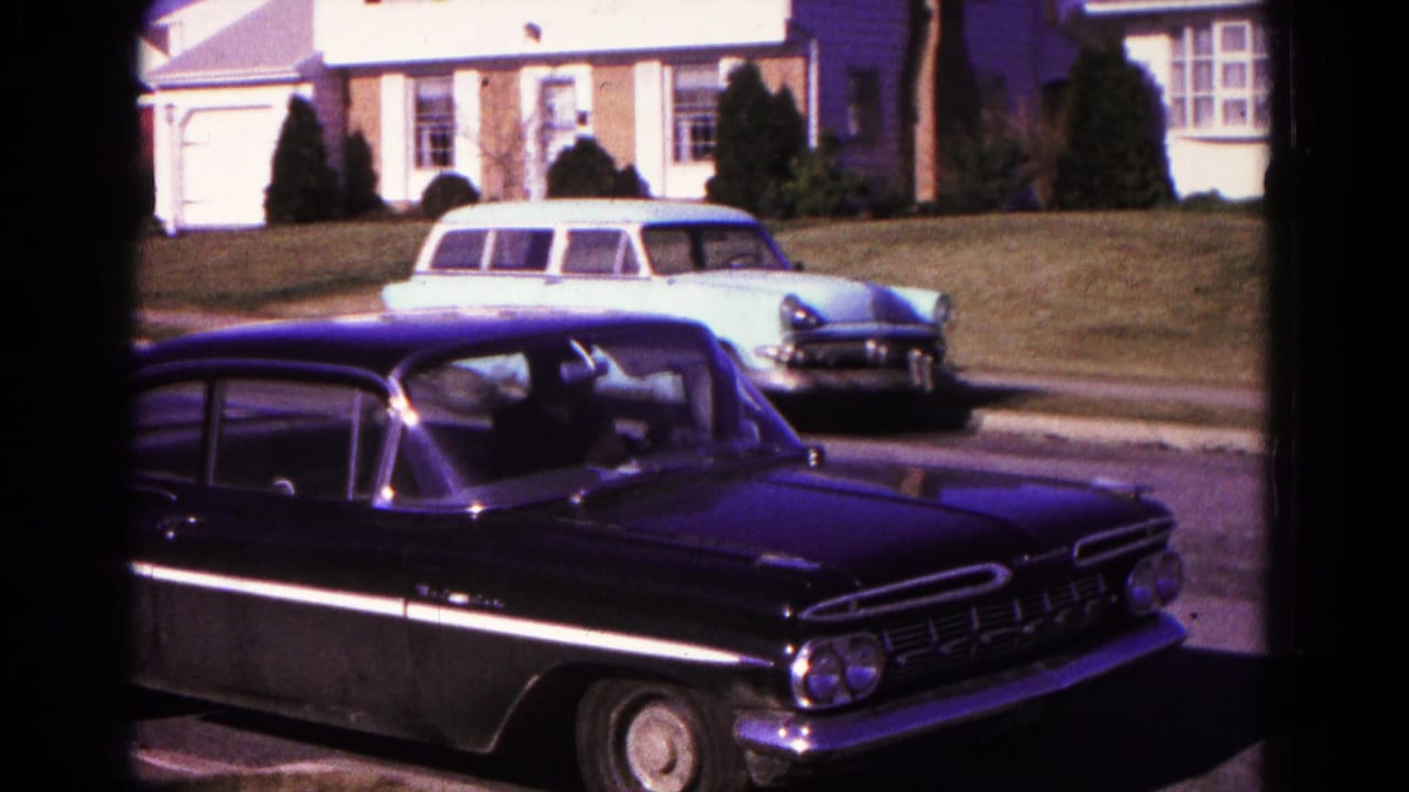 Black sedan in the foreground with a light blue station wagon parked near a suburban house.
