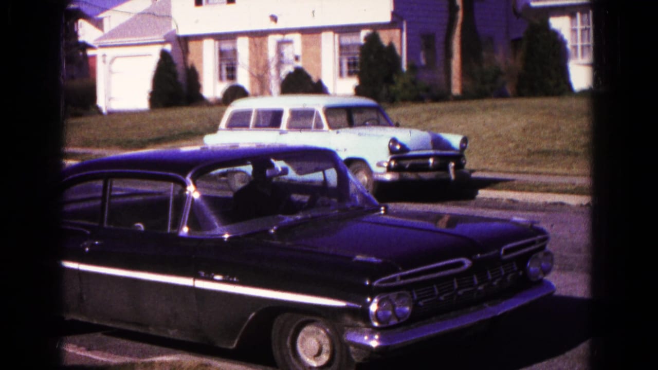Black sedan moving across the foreground while a station wagon remains parked in front of a house.