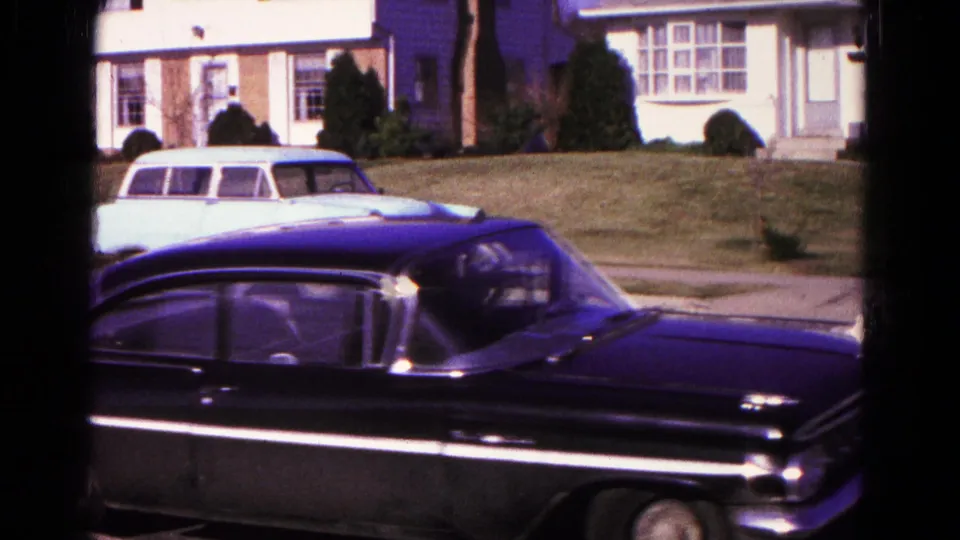 Close 8mm frame of the black sedan passing through a Hagerstown neighborhood, with a station wagon, lawn, and house visible behind it.