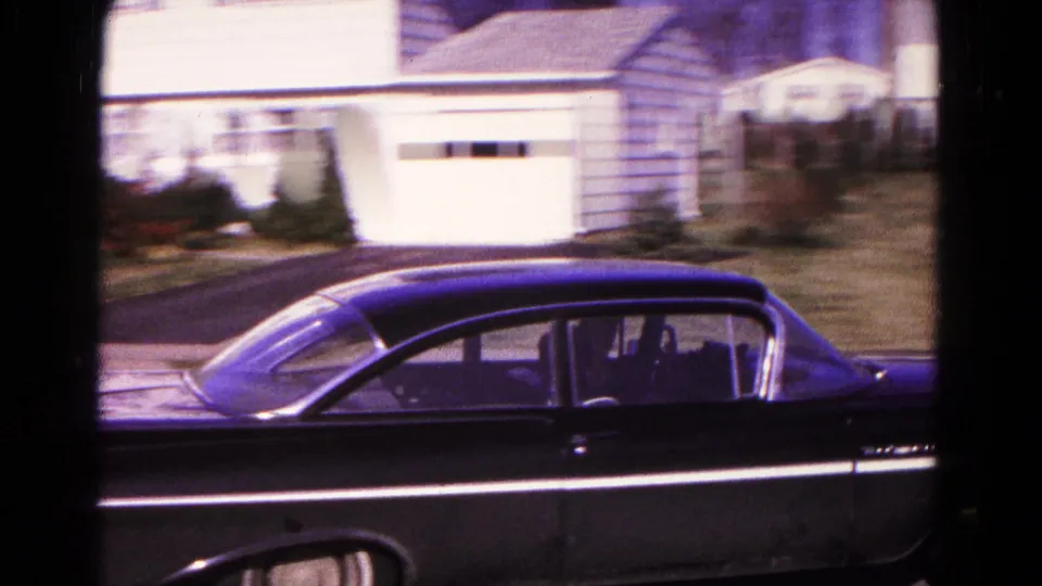 Side view of the black sedan near a driveway and garage in 1966 Hagerstown, with handheld motion blur across the residential background.