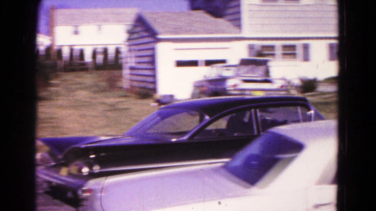 Black classic sedan passing a white car on a suburban street beside detached houses.