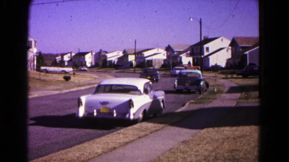 A white classic car remains near the curb in a 1966 Hagerstown neighborhood as another car continues down the street.