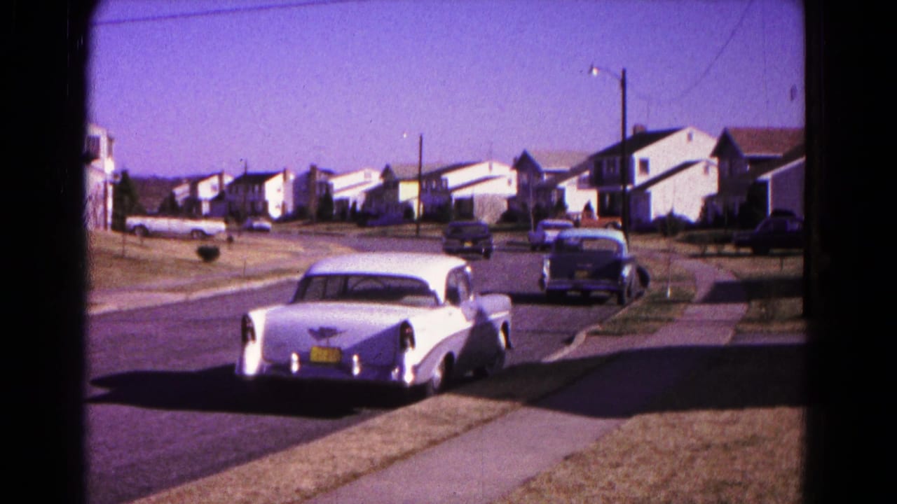 White mid-century car on a sunny residential street with detached houses and parked cars.
