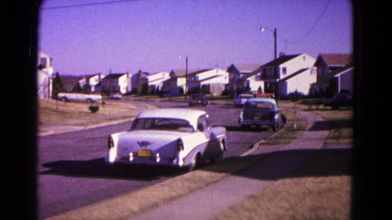 Sunny suburban street with a white car in the foreground and houses in the distance.