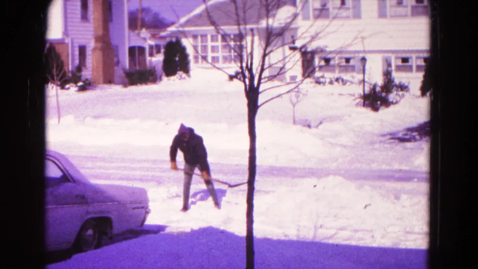 1966 Hagerstown winter home-movie frame showing a man shoveling snow near a parked car, bare tree, and snow-covered residential lawns.