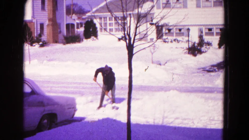 1966 Hagerstown winter scene with a man shoveling heavy snow beside a parked car, a bare tree, and detached suburban houses.