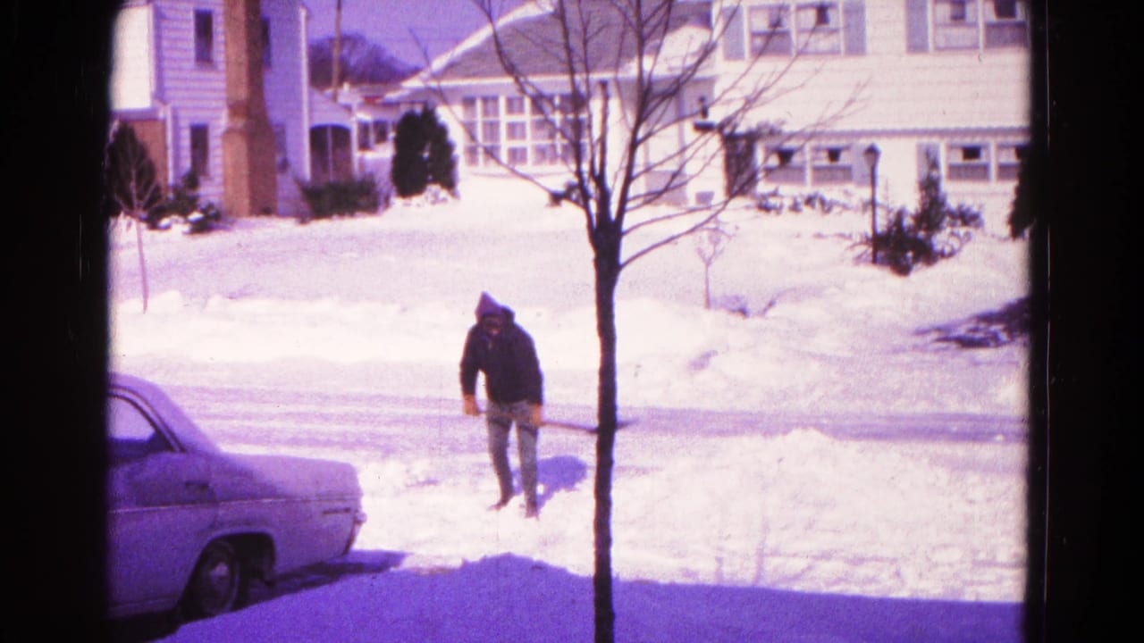 Man standing with a shovel near a snowbank beside a parked sedan on a suburban street.