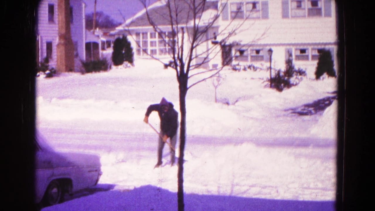 Man bends over a shovel while clearing snow near a driveway and parked car.